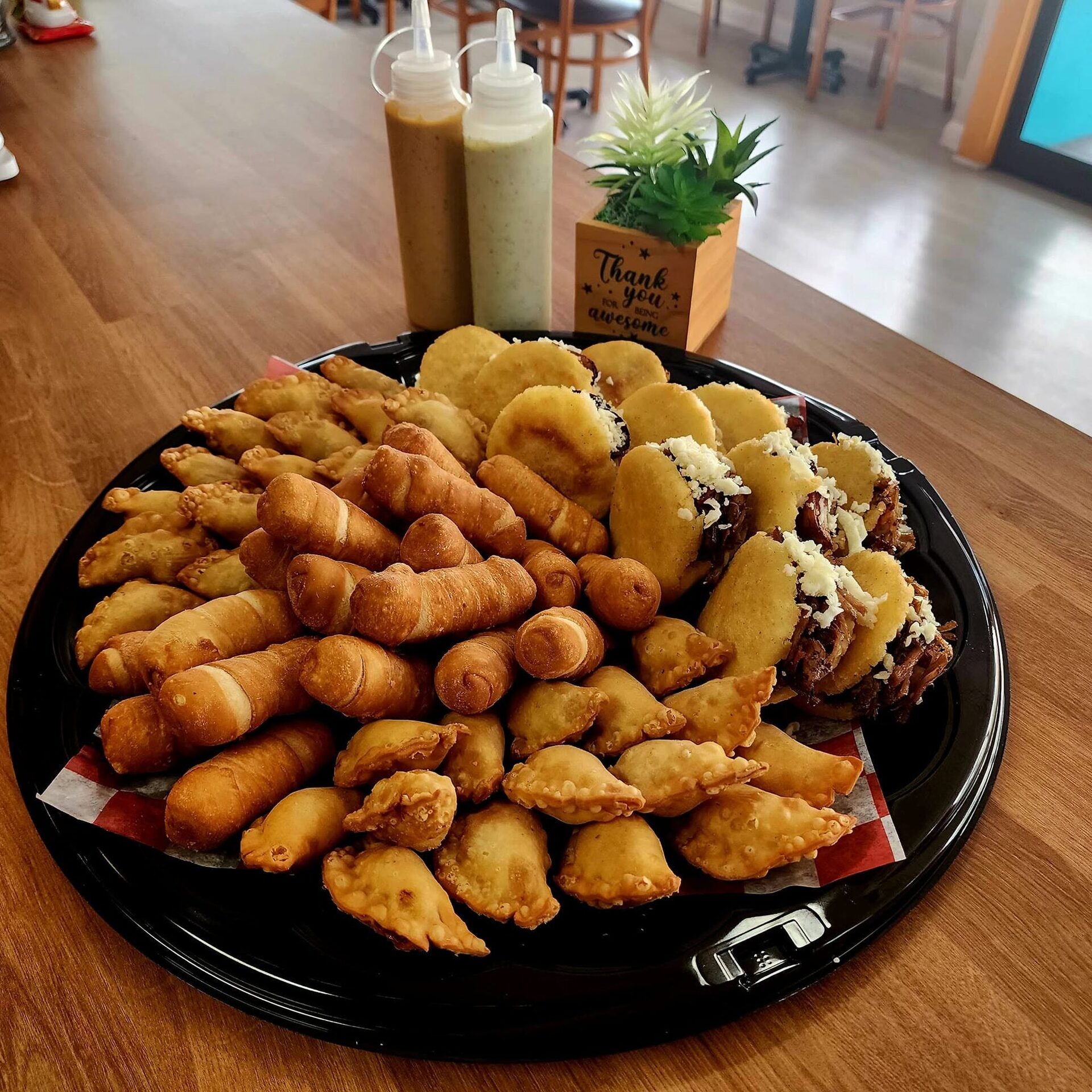 A loaded 1Chick 1Bro platter of empanadas, tequeños and mini arepas with sauce bottles behind