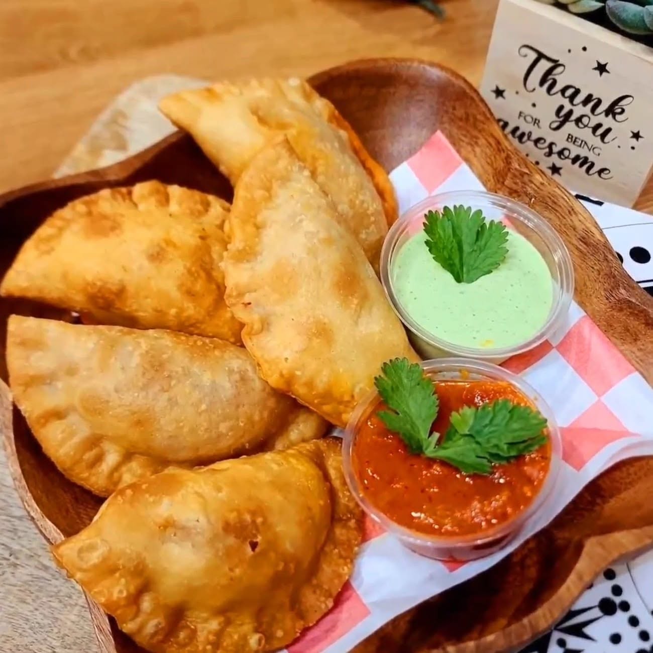 Hand-folded empanadas with the house green sauce and a red sauce in a wooden bowl