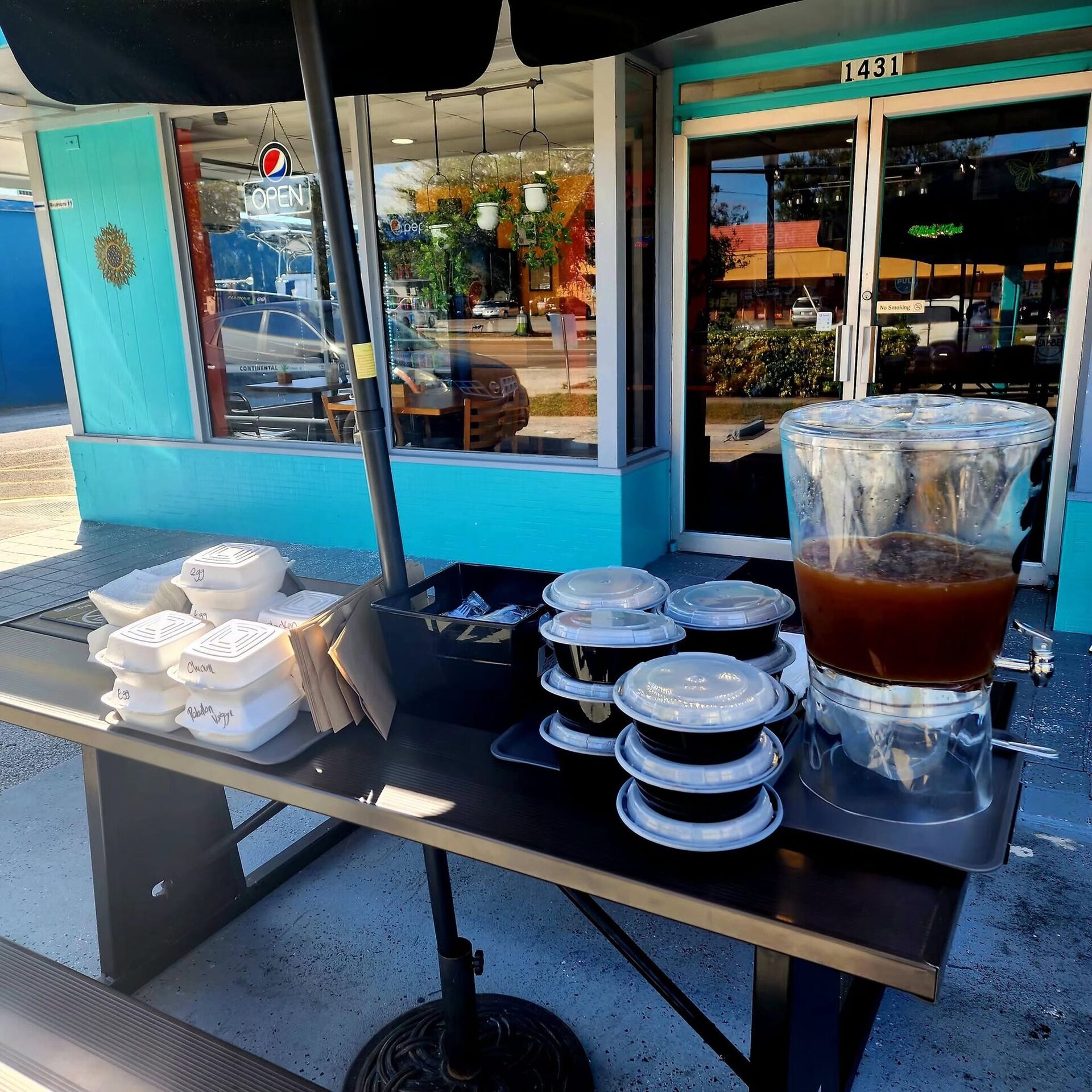 1Chick 1Bro to-go trays and a drink dispenser set out on the front patio table by the teal storefront