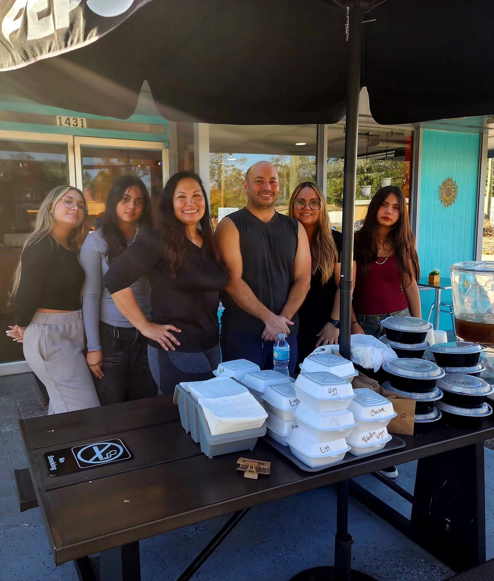 The 1Chick 1Bro family standing in front of the store behind a table of packed catering trays